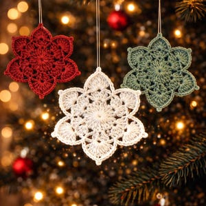 May include: Three crocheted snowflake decorations in red, white, and green hang against a blurred Christmas tree backdrop. The ornaments, with a six-petal design and intricate detailing, are ideal for festive decor.