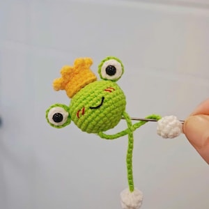 May include: A handcrafted green crochet frog wearing a yellow crown. The frog has large, round eyes with black pupils, a smiling mouth, and small red cheeks. It has white pom-poms at the end of its arms and legs.