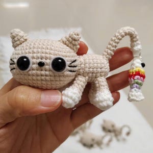 Op de afbeelding: Een gehaakte kattenfiguur in beige met grote zwarte ogen en witte poten. De kat houdt een kleurrijke vissenbedel vast. De staart van de kat is gebogen en de vissenbedel heeft rode, gele, groene en paarse details.
