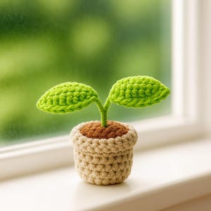 Könnte beinhalten: Gehäkelte Pflanze in einem kleinen Topf. Die Pflanze hat zwei leuchtend grüne Blätter und eine braune Basis, in einem beigefarbenen Topf. Der Topf steht auf einer weißen Fensterbank mit verschwommenem grünem Hintergrund.