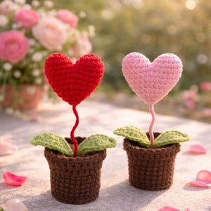 May include: Two crochet heart-shaped flowers in brown pots. One heart is red, the other is pink. Green leaves and stems support the hearts. The pots sit on a stone surface with a blurred background of flowers.