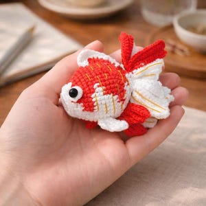 Könnte beinhalten: Handgehäkelter Goldfisch in Rot, Weiß und Gold. Der Fisch hat einen runden Körper, große schwarze Augen und fließende Flossen. Der Fisch wird in einer Hand gehalten, was die komplizierten Details des Handwerks zeigt.