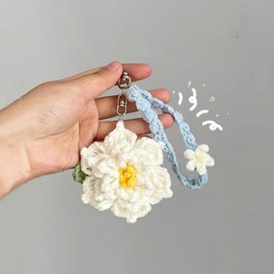 May include: A handmade crochet keychain featuring a large white flower with a yellow center and a small green leaf. The keychain has a light blue braided strap and a silver-colored clasp.