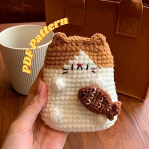 May include: A crochet cat-shaped case in white and brown, holding a brown fish. The cat has a red nose and black eyes and whiskers. The text "PDF Pattern" is visible in the background.