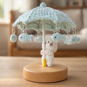 May include: A handmade crochet mobile featuring a white character holding a light blue umbrella. The umbrella has dangling blue fish and is mounted on a wooden base. The mobile is a decorative item.