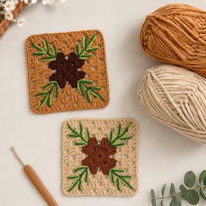 May include: Two square crochet coasters featuring a central dark brown flower motif surrounded by green embroidered pine needle sprigs. One coaster is tan and the other is beige, accompanied by two matching balls of yarn and a wooden crochet hook.