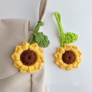 May include: Two handmade crochet sunflower charms. The sunflowers have yellow petals and brown centers. Green stems and leaves are attached. One charm is attached to a beige bag. The other charm is hanging freely.