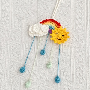 May include: A handmade crochet mobile featuring a rainbow, fluffy clouds, and a smiling sun. The mobile has blue and green teardrop-shaped accents hanging from the clouds and sun. The rainbow has red, orange, yellow, green, blue, and purple colors.