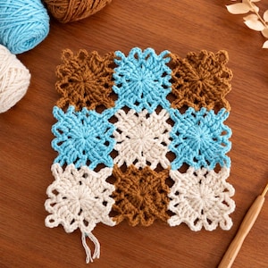 May include: A square crochet piece composed of nine individual motifs arranged in a three-by-three grid. The motifs feature a starburst pattern in alternating colors of light blue, cream, and brown, with scalloped edges and visible yarn ends.