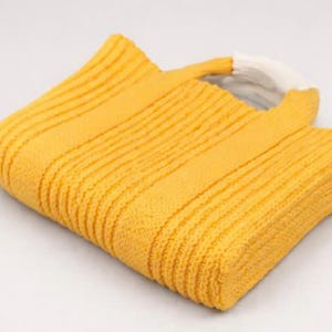 May include: A yellow knitted tote bag with a white handle. The bag is made of a textured, ribbed fabric. The bag is folded over, showing the interior. The bag is on a white background.