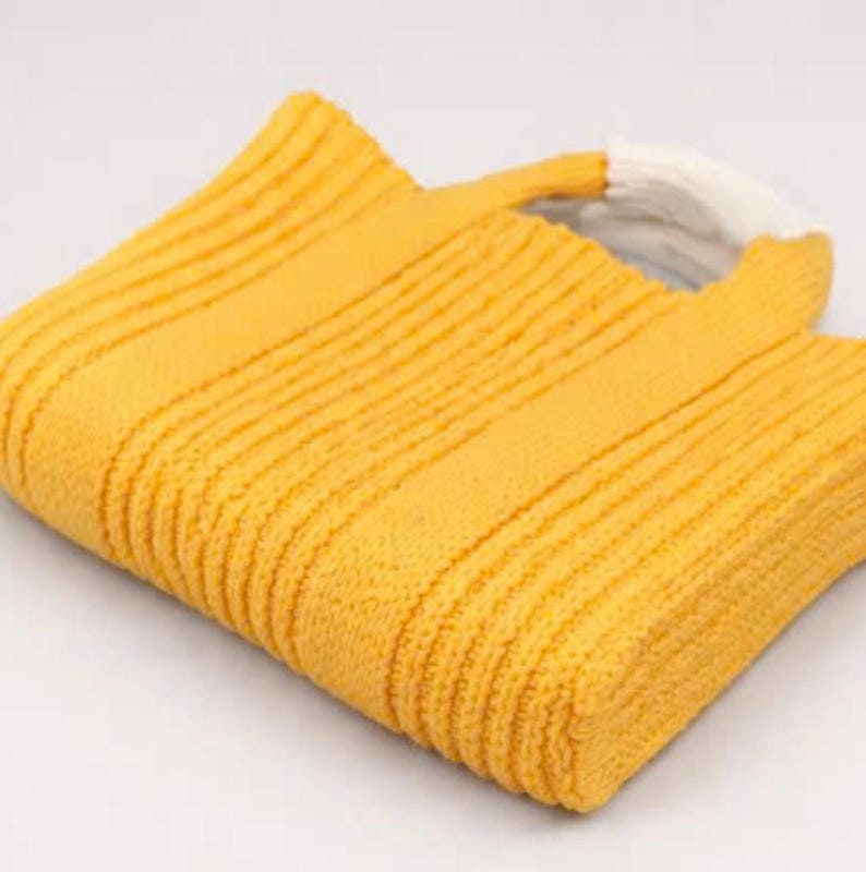 May include: A yellow knitted tote bag with a white handle. The bag is made of a textured, ribbed fabric. The bag is folded over, showing the interior. The bag is on a white background.