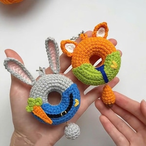 Puede incluir: Dos llaveros de ganchillo con forma de rosquilla. Uno gris con orejas de conejo y una zanahoria, el otro naranja con orejas de zorro y una zanahoria. El llavero de conejo tiene un diseño azul y gris, y el llavero de zorro tiene un diseño verde y naranja.