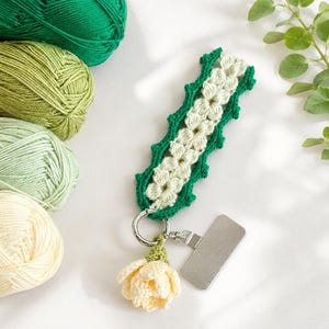 May include: A handmade crochet keychain featuring a green and white floral design. The keychain has a silver clasp and a small, cream-colored crochet flower. Green and cream-colored yarn balls are in the background.