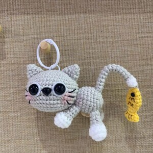 Op de afbeelding: Een gehaakte grijze katten sleutelhanger met grote zwarte ogen, roze wangen en witte poten. De kat houdt een gele vis vast. De sleutelhanger heeft een witte ring om aan te bevestigen.