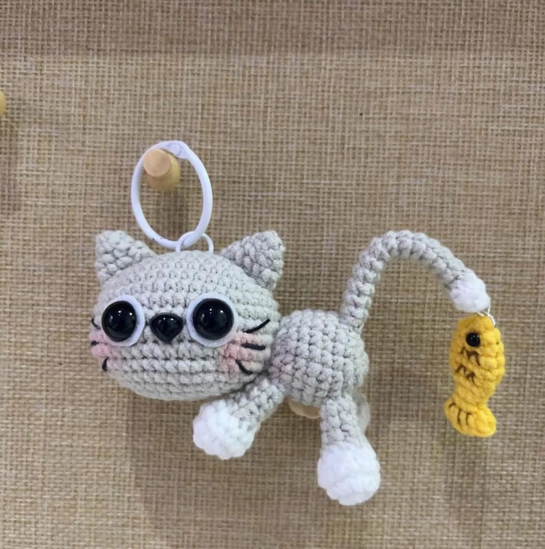 Op de afbeelding: Een gehaakte grijze katten sleutelhanger met grote zwarte ogen, roze wangen en witte poten. De kat houdt een gele vis vast. De sleutelhanger heeft een witte ring om aan te bevestigen.