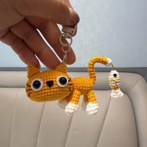 Op de afbeelding: Een handgemaakte oranje gehaakte katten sleutelhanger met witte poten en een witgepunte staart. De kat houdt een kleine gehaakte vis vast. De sleutelhanger heeft een metalen sluiting.