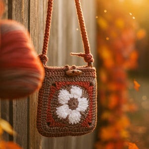 Op de afbeelding: Handgemaakte gehaakte tas met een bloemenpatroon. De tas heeft een bruin, oranje en wit kleurenschema, met een houten knevelsluiting en een gevlochten band. De tas hangt aan een houten oppervlak.