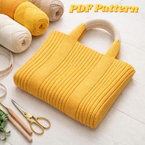 May include: A mustard yellow knitted tote bag with cream handles, with yarn and craft tools. The bag has a ribbed texture and the text "PDF Pattern" is visible. Gold scissors and crochet hooks are also present.