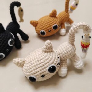 Op de afbeelding: Gehaakte kattenbeeldjes in zwart, bruin en cr&egrave;me. Elke kat heeft grote zwarte ogen en een gebogen staart. Sommige katten houden een kleine vis vast. De handgemaakte speelgoedjes zijn geschikt voor kinderen of als decoratie.