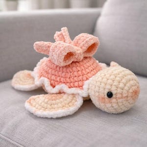 May include: A handmade crochet turtle plushie in soft peach and cream colors. The turtle has a peach shell with a white trim, cream-colored flippers, and a cream head with a black eye. A peach bow adorns the turtle's back.