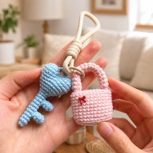 Può includere: Un portachiavi all'uncinetto fatto a mano con una chiave azzurra e un lucchetto rosa, entrambi con disegni a forma di cuore. La chiave e il lucchetto sono collegati da un anello di metallo e un anello di corda beige.