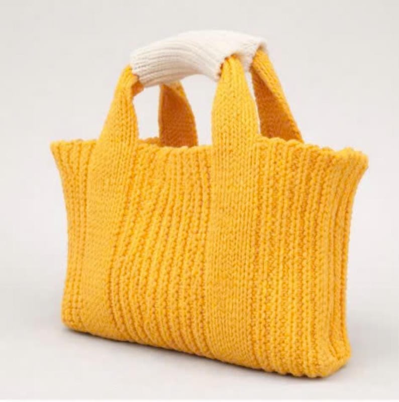 May include: A mustard yellow knitted tote bag with a cream-coloured handle. The bag has a rectangular shape with vertical ribbing and two short handles. The handle is a thick, knitted band.