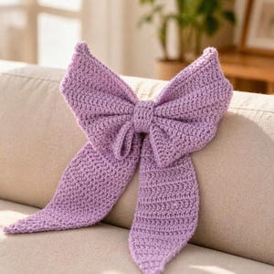 May include: A large, lavender-colored crochet bow. The bow is made of a soft, textured yarn and has long, flowing tails. The bow is sitting on a beige couch.