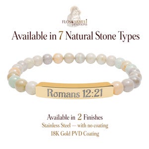 May include: A stretch bracelet featuring smooth, multi-colored natural stone beads in shades of beige, grey, and pale green. A central gold-toned metal bar is engraved with the text Romans 12:21. Text above reads Flos Carmeli Devotions.