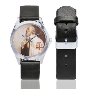 May include: A silver wristwatch with a black leather band. The watch face features a portrait of a man in religious attire. The man has a white beard and is wearing a white robe with a red cross emblem. The watch band is detached.