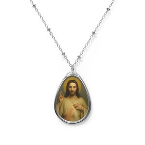 May include: Silver-toned pendant necklace featuring an oval pendant with a religious image. The pendant depicts a figure with long hair, a halo, and a hand raised in blessing. The chain is a delicate link style.