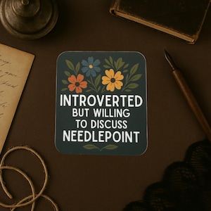 May include: A square, dark green coaster with the text "INTROVERTED BUT WILLING TO DISCUSS NEEDLEPOINT" in white. The coaster features floral designs in orange, yellow, and blue.
