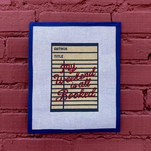 May include: A framed needlepoint piece with a blue border against a red brick wall. The artwork features a cream-colored background with a design resembling a library card. The text "My Weekend is all Booked" is in red cursive.