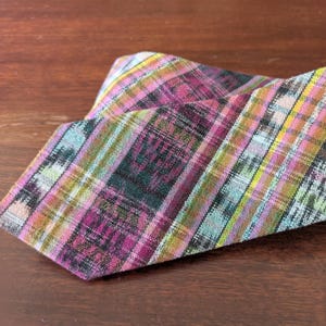 May include: A colorful plaid necktie with a mix of purple, green, yellow, and blue hues. The tie is made of a woven fabric and is displayed on a wooden surface. The diagonal pattern adds visual interest.
