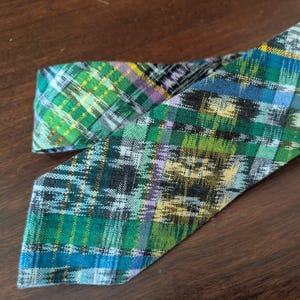 May include: A colorful necktie with a plaid pattern in shades of green, blue, yellow, and black. The tie is made of fabric and is folded over itself. The tie is laying on a dark brown wooden surface.