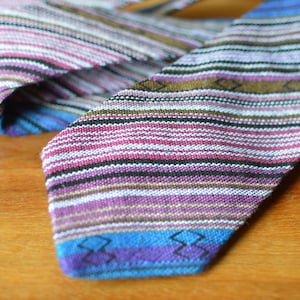 May include: A colorful necktie with a striped pattern in shades of purple, pink, blue, and brown. The tie is made of woven fabric and has a geometric design at the tip. The tie is lying on a wooden surface.