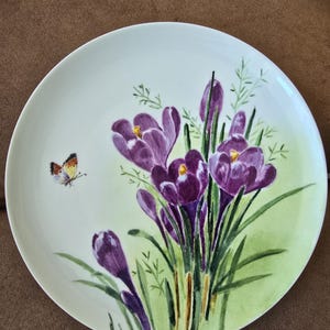 May include: A white ceramic plate featuring a hand-painted floral design. The design includes purple crocus flowers with yellow centers, green leaves, and a small butterfly. The plate has a light green background.