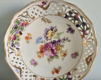 Peut inclure: Une assiette en porcelaine blanche avec un motif floral. L'assiette présente un bord doré et une bordure ajourée. Le centre de l'assiette est orné d'un bouquet de fleurs violettes, jaunes, oranges et roses.