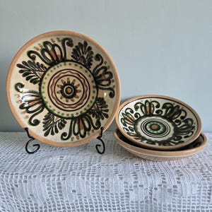 Könnte beinhalten: Ein Satz dekorativer Keramikteller mit beigem Rand und cremefarbenem Zentrum. Die Teller weisen ein florales Design in Braun-, Grün- und Orangetönen auf. Der größte Teller wird auf einem schwarzen Metallständer präsentiert.