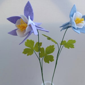 May include: Two felt flowers in a white vase. The flowers are purple and blue with white centers and yellow stamens. The stems are green and the leaves are green.