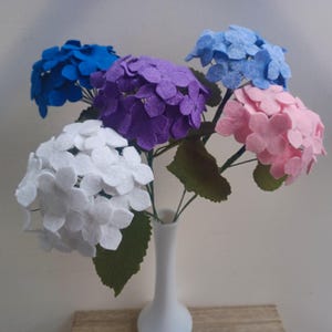 May include: A bouquet of five felt hydrangea flowers in a white vase. The flowers are white, pink, purple, and blue.