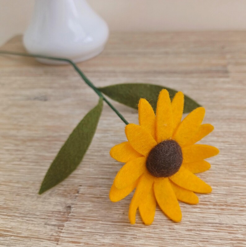 Op de afbeelding: Een enkele viltzonnebloem met een bruin centrum en groene bladeren bevestigd aan een draadsteel. De zonnebloem is geel en de bladeren zijn donkergroen.
