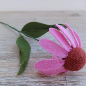 May include: A handmade felt flower with a pink bloom and a brown center. The flower has green leaves and a green stem. The flower is lying on a light-colored wooden surface.