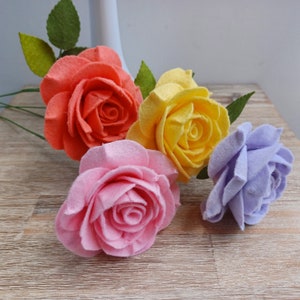 May include: Four felt roses in different colors: orange, yellow, pink, and lavender. The roses are arranged on a wooden surface.