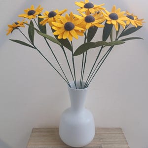 Op de afbeelding: Een boeket van viltzonnebloemen in een witte vaas. De zonnebloemen zijn geel met bruine centra en groene bladeren. De vaas is een eenvoudige, witte vaas met een ronde bodem.