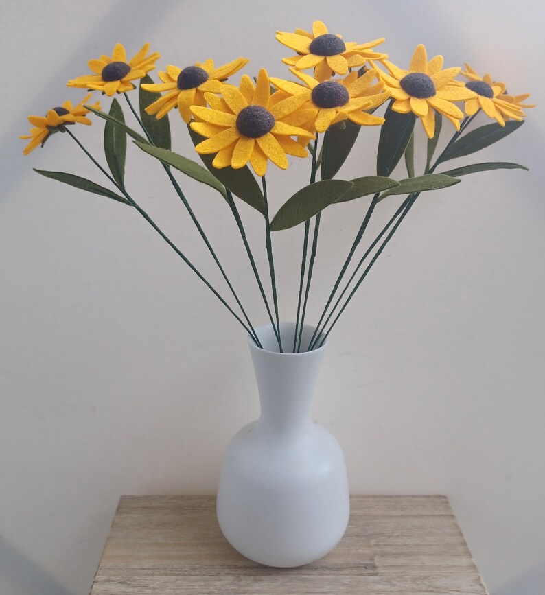 Op de afbeelding: Een boeket van viltzonnebloemen in een witte vaas. De zonnebloemen zijn geel met bruine centra en groene bladeren. De vaas is een eenvoudige, witte vaas met een ronde bodem.