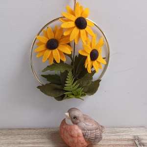 Puede incluir: Un aro de metal dorado con tres girasoles de fieltro amarillo y hojas de fieltro verde. Una peque&ntilde;a figurita de p&aacute;jaro de cer&aacute;mica marr&oacute;n y naranja est&aacute; en primer plano.