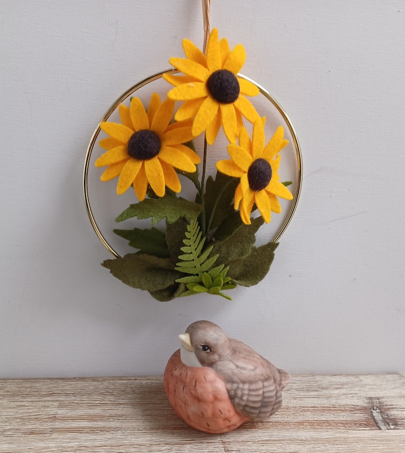 Puede incluir: Un aro de metal dorado con tres girasoles de fieltro amarillo y hojas de fieltro verde. Una peque&ntilde;a figurita de p&aacute;jaro de cer&aacute;mica marr&oacute;n y naranja est&aacute; en primer plano.