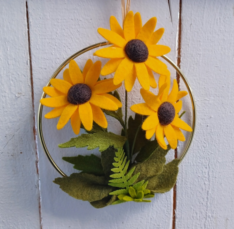 Puede incluir: Un aro de metal dorado con tres girasoles de fieltro. Los girasoles son amarillos con centros marrones y hojas verdes.