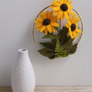 Puede incluir: Un aro de metal dorado con tres girasoles amarillos de fieltro y hojas verdes de fieltro. Un jarr&oacute;n de cer&aacute;mica blanco con superficie texturizada est&aacute; en primer plano.