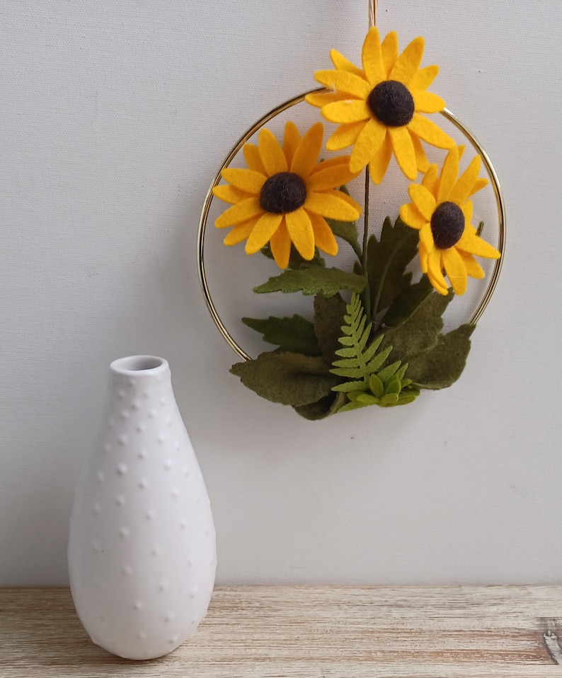 Puede incluir: Un aro de metal dorado con tres girasoles amarillos de fieltro y hojas verdes de fieltro. Un jarr&oacute;n de cer&aacute;mica blanco con superficie texturizada est&aacute; en primer plano.